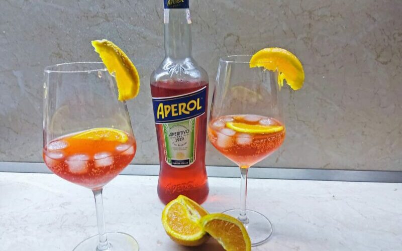 Fľaša s Aperolom a dva poháre s drinkom Aperol Spritz ozdobené pomarančom