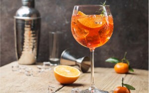 Aperol Spritz koktail - na stole v pohári v pozadí so šejkrom a rozkrojeným pomarančom