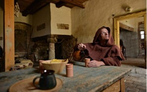 Stredoveký škótsky mních - sedí za stolom a nalieva si do pohára whisky (nejaký alkoholický nápoj)