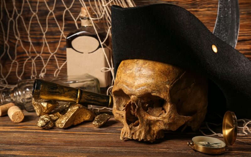 Piráti a rum: Ľudská lebka s pirátskym klobúkom, zlatými nugetami, fľašou rumu a cestovným vybavením na hnedom dreve